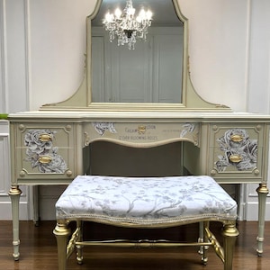 May include: A sage green vanity table with an arched mirror, gold-toned hardware, and floral decals. The center drawer features the text "CREAM COLLECTION OF 12 EVER BLOOMING ROSES". A matching bench with a white and gray floral fabric seat is included.
