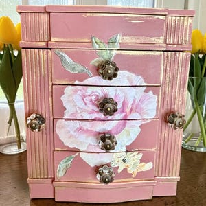 May include: A pink jewelry box with four drawers, adorned with a large floral design and bronze-colored knobs. The box has gold accents and a distressed finish, giving it a vintage aesthetic. The box is a decorative storage solution.