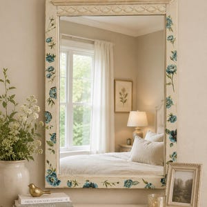 May include: A rectangular wall mirror with a cream-colored frame featuring hand-painted blue floral motifs. The frame has a decorative arched top and a carved horizontal molding detail. The mirror reflects a bedroom interior with a bed and window.