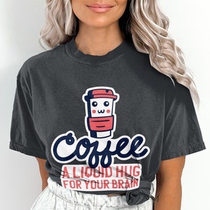 Może przedstawiać: Ciemnoszara koszulka z grafiką filiżanki kawy w kolorze czerwonym i białym. Tekst "Coffee, A Liquid Hug For Your Brain" jest napisany zabawną czcionką. Dla miłośników kawy.