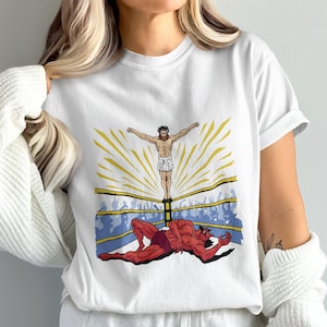 Könnte beinhalten: Weißes T-Shirt mit einem Grafikdruck eines Mannes in einem Wrestling-Ring. Der Mann steht auf einer Plattform mit einem Heiligenschein über seinem Kopf. Ein roter Teufel liegt auf dem Boden im Ring.