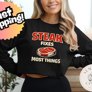 Puede incluir: Top corto negro de manga larga con el texto "STEAK FIXES MOST THINGS" en rojo y blanco. El gráfico incluye una ilustración de bistec. Una pegatina rosa y amarilla de "Fast Shipping!" está en la esquina superior izquierda.