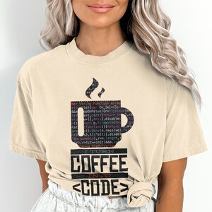 May include: Beige t-shirt with a coffee cup graphic filled with computer code and steam. Below the cup, the text reads "I TURN COFFEE INTO CODE" in a bold, black font. The shirt is a comfortable, casual style.