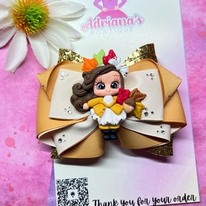 May include: A handmade hair bow featuring a detailed figurine of a girl in a yellow dress, holding acorns. The bow is a combination of tan and cream-colored ribbons, accented with gold glitter and rhinestones. The card reads "Adriana's Boutique" and "Thank you for your order."