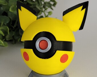 Könnte beinhalten: Ein gelber und schwarzer Pokéball mit Pikachu-Ohren. Der Pokéball hat einen roten Knopf und einen schwarzen Ring um die Mitte. Der Pokéball steht auf einem grauen Ständer.