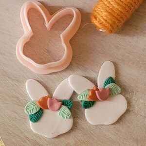 May include: Two white bunny shaped polymer clay earrings with green leaves and orange and pink flower accents. A pink bunny shaped cookie cutter is also pictured.