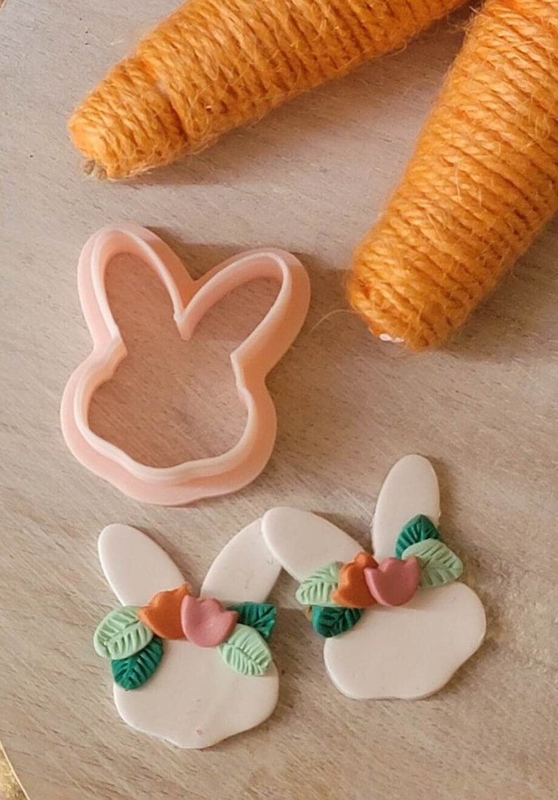 May include: Two white bunny shaped polymer clay earrings with green leaves and orange and pink flower accents. A pink bunny shaped cookie cutter is also pictured.