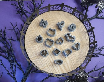 Puede incluir: Un juego de 14 cortadores de galletas de Halloween de plástico gris en una bandeja de madera. Los cortadores tienen forma de calabaza, fantasma, murciélago, sombrero de bruja, ataúd, telaraña y otras formas con temática de Halloween.