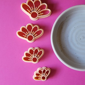 Op de afbeelding: Vier verschillende maten plastic koekjesvormen in de vorm van een bloem met een rood midden en oranje bloemblaadjes. De vormen zijn verticaal gerangschikt op een roze oppervlak.