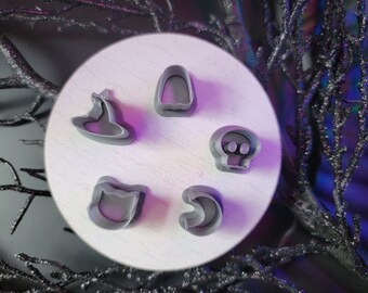 May include: Five gray Halloween-themed cookie cutters arranged on a white surface. The shapes include a witch hat, a tombstone, a skull, a cat face, and a crescent moon. The background features dark, glittery branches.
