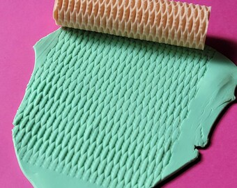 May include: A light green rolling pin with a textured surface is being used to create a pattern in a piece of light green clay.