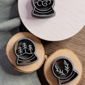 May include: Three gray metal cookie cutters shaped like snow globes. The first snow globe has a crescent moon and clouds inside. The second snow globe has three pine trees inside. The third snow globe has a sprig of holly inside.