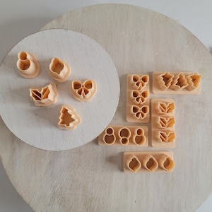 Könnte beinhalten: Ein Set mit 13 Ausstechern aus Kunststoff in verschiedenen Formen, darunter Schleifen, Blätter, ein Weihnachtsbaum und Kreise. Die Ausstecher sind beige und liegen auf einer hellbraunen Holzoberfläche.