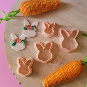 May include: A set of four pink bunny cookie cutters and two handmade bunny decorations with green leaves and pink flowers. The decorations are on a wooden surface with two orange yarn carrots.