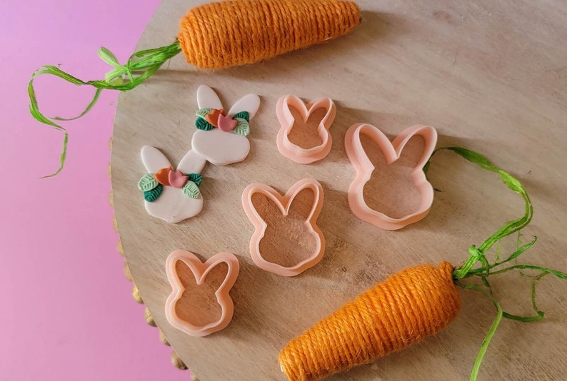 May include: A set of four pink bunny cookie cutters and two handmade bunny decorations with green leaves and pink flowers. The decorations are on a wooden surface with two orange yarn carrots.