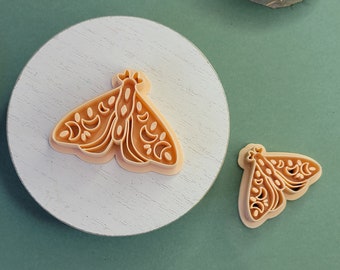 May include: Two tan-colored moth-shaped cookie cutters. One is larger and rests on a white, round surface. The other is smaller and sits on a green surface. Both cutters have intricate wing and body details.