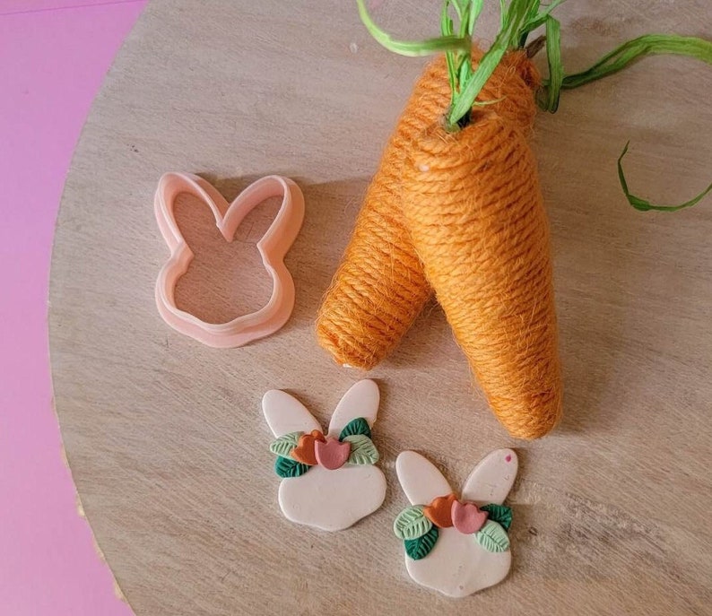 May include: Two white bunny shaped clay ornaments with green and pink floral accents. The ornaments are sitting on a wooden surface next to two yarn wrapped carrots.