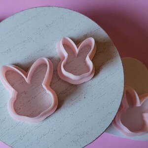 May include: Three pink plastic bunny cookie cutters on a white wooden surface. The cutters are different sizes and shapes.