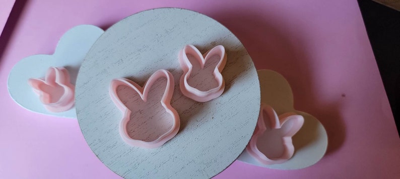 May include: Three pink plastic bunny cookie cutters on a white wooden surface. The cutters are different sizes and shapes.