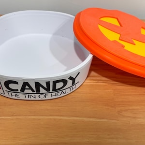 May include: A white round container with a small grey handle and a removable orange lid featuring a yellow cutout design. The side of the container displays a red cross symbol and the black text CANDY THE TIN OF HEALTH in a bold font.