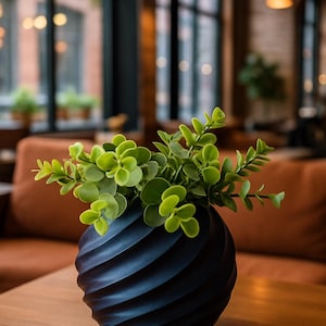 May include: A dark blue vase with a spiral design, filled with green foliage. The vase is on a wooden table. The background shows a room with windows and seating. The vase is a decorative item.