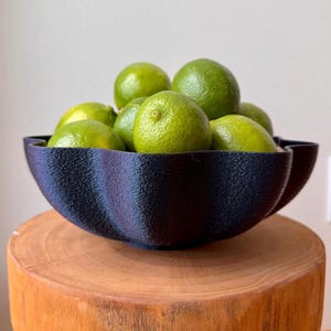May include: A dark blue decorative bowl filled with bright green limes. The bowl has a textured surface and sits on a wooden stump. The limes are round and have a smooth skin. The bowl's design is wavy, adding a unique touch to the fruit display.