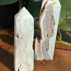 May include: Two carved stone obelisks. The obelisks are light green with tan and brown veining. One is intact, while the other is split, revealing a hollow interior. The obelisks are displayed on a wooden surface.