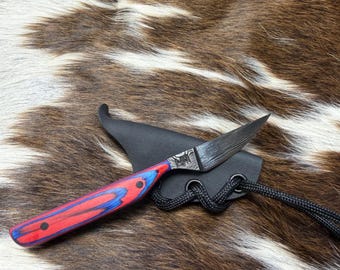 May include: A small, handcrafted knife with a patterned blade and a red and blue handle. The knife rests in a black leather sheath with a black cord. The background is a brown and white animal hide.