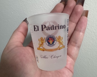 Pode incluir: Um copo de shot de vidro transparente com o texto "El Padrino" e um logotipo da Cerveceria Modelo Mexico 1925. O logotipo apresenta um leão e uma coroa. O texto "Mas Chingon" está impresso abaixo do logotipo.