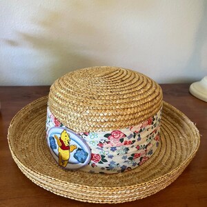 May include: A straw hat with a wide brim and a rounded crown. The hat features a floral patterned band with a Winnie the Pooh patch. The hat is made of woven straw and is suitable for sun protection.