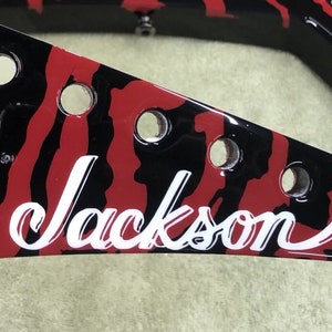 May include: Close-up of a guitar headstock with a red and black zebra stripe pattern. The word "Jackson" is written in white script. The headstock has six circular holes.