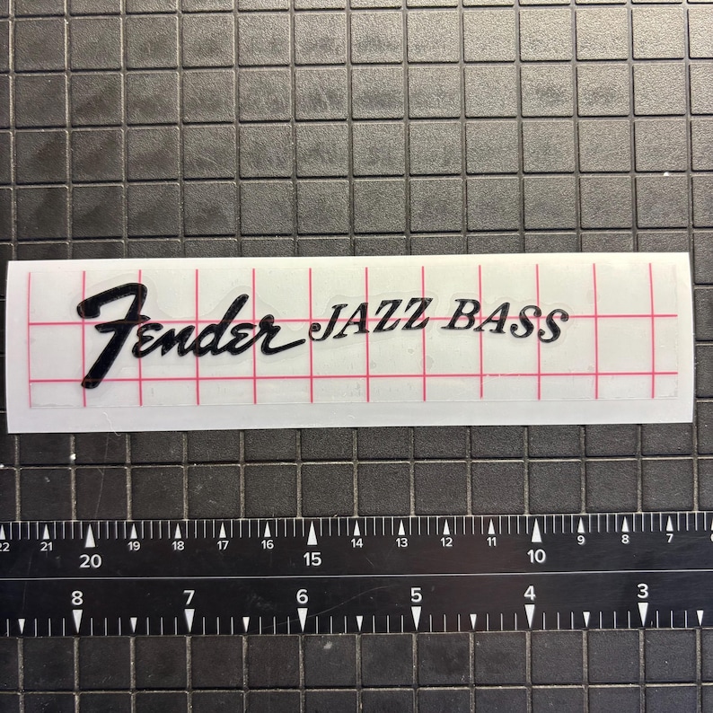 May include: A transparent sticker featuring the text "Fender JAZZ BASS" in black script and block letters. The sticker is on a white background with a red grid. A ruler is below the sticker.