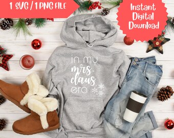 Op de afbeelding: Een grijze hoodie met de tekst "In My Mrs. Claus Era" in witte letters en sneeuwvlokken. De hoodie ligt op een wit houten oppervlak met een paar bruine laarzen met nep bontvoering, een blauwe jeans, een koffiemok en kerstversiering.