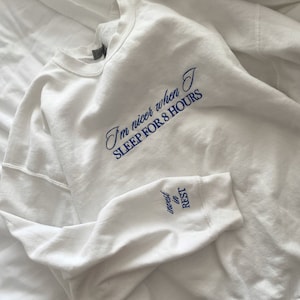 Peut inclure: Sweat-shirt blanc avec un texte brodé en bleu qui dit "Je suis plus gentil quand je dors 8 heures" et "investir dans le repos" sur la manche.