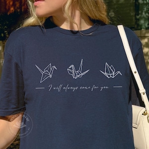 May include: A navy blue t-shirt with a white origami crane design and the text "I will always come for you". The design is simple and stylish, perfect for everyday wear.