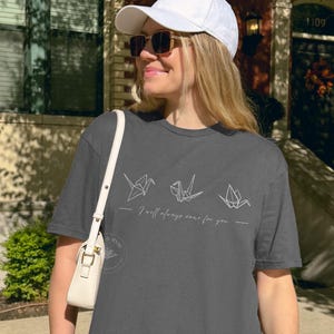 May include: A dark grey t-shirt with origami crane designs and the phrase "I will always care for you." The outfit includes a white baseball cap and sunglasses. A white handbag is also visible.