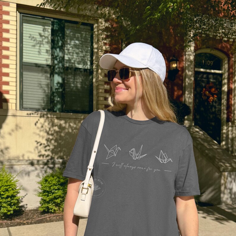 May include: A dark grey t-shirt with origami crane designs and the phrase "I will always care for you." The outfit includes a white baseball cap and sunglasses. A white handbag is also visible.