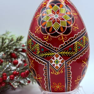 May include: A hand-painted decorative egg with a deep red base and detailed floral patterns in yellow, orange, black, and white. The egg is displayed on a clear stand, highlighting the folk art design. Greenery with red berries is in the background.