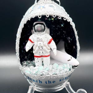 May include: An egg-shaped ornament with an astronaut and a white airplane inside. The ornament is white with silver trim and the words "Reach the stars". The interior has a starry night design. The ornament sits on a clear stand.