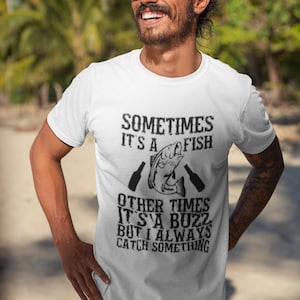 Puede incluir: Camiseta blanca con un gráfico negro de un pez y texto que dice "Sometimes it's a fish other times it's a buzz but I always catch something."