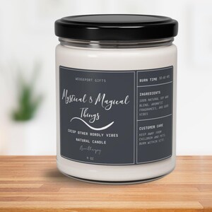 Op de afbeelding: Een witte kaars in een glazen pot met een zwarte deksel. Het etiket is donkergrijs met witte tekst die "Mystical & Magical Things", "Crisp Other Wordly Vibes", "Natural Candle" en "Burn Time 50-60 hrs" luidt.