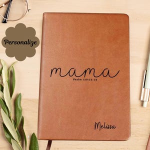 Puede incluir: Un diario de cuero marrón con la palabra "mama" en escritura cursiva y el nombre "Melissa" debajo. El diario también tiene el texto "Salmo 130:13-14". Un bolígrafo y gafas están sobre una superficie de madera.