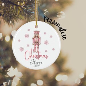 May include: A white round Christmas ornament with a pink watercolor illustration of a nutcracker, snowflakes, and the text "1st Christmas Olivia 2024".