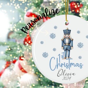 May include: White ceramic Christmas ornament with a blue and white illustration of a nutcracker, snowflakes, and the text "1st Christmas Olivia 2024".