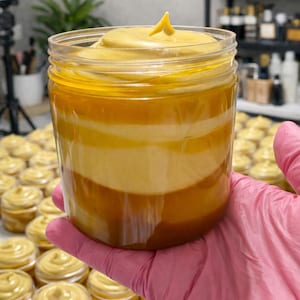 May include: A clear plastic jar filled with a thick, layered cream featuring distinct bands of golden yellow, cream, and deep amber colors. The top of the cream is swirled into a peak, and the jar is held in a hand wearing a pink glove.