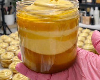 May include: A clear plastic jar filled with a thick, layered cream featuring distinct bands of golden yellow, cream, and deep amber colours. The top of the cream is swirled into a peak, and the jar is held in a hand wearing a pink glove.