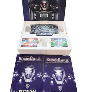 May include: A black and silver handheld video game console called "Barcode Battler" with two game cartridges and instruction manuals. The game console has a screen that displays the text "Card Battle Setup". The box for the game console features a robot with red eyes and the text "Commerce Conflict Barcode Battler".