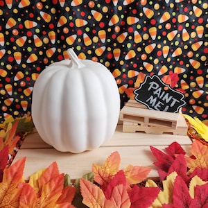 Puede incluir: Una gran calabaza blanca sin pintar con una superficie texturizada, lista para pintar. Se encuentra sobre una paleta de madera con un pequeño letrero de pizarra que dice "Paint Me!". El fondo presenta un patrón de caramelos.