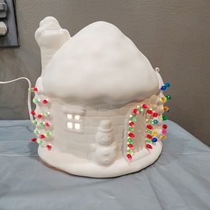 May include: White ceramic Christmas house with a snowy roof and a lit window. The house is decorated with colorful string lights.