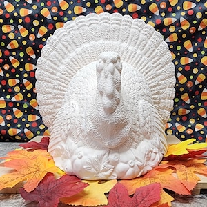 Puede incluir: Figurita de pavo de cerámica blanca con una cola de plumas detallada y un cuerpo texturizado. El pavo está sentado en un lecho de hojas de otoño.